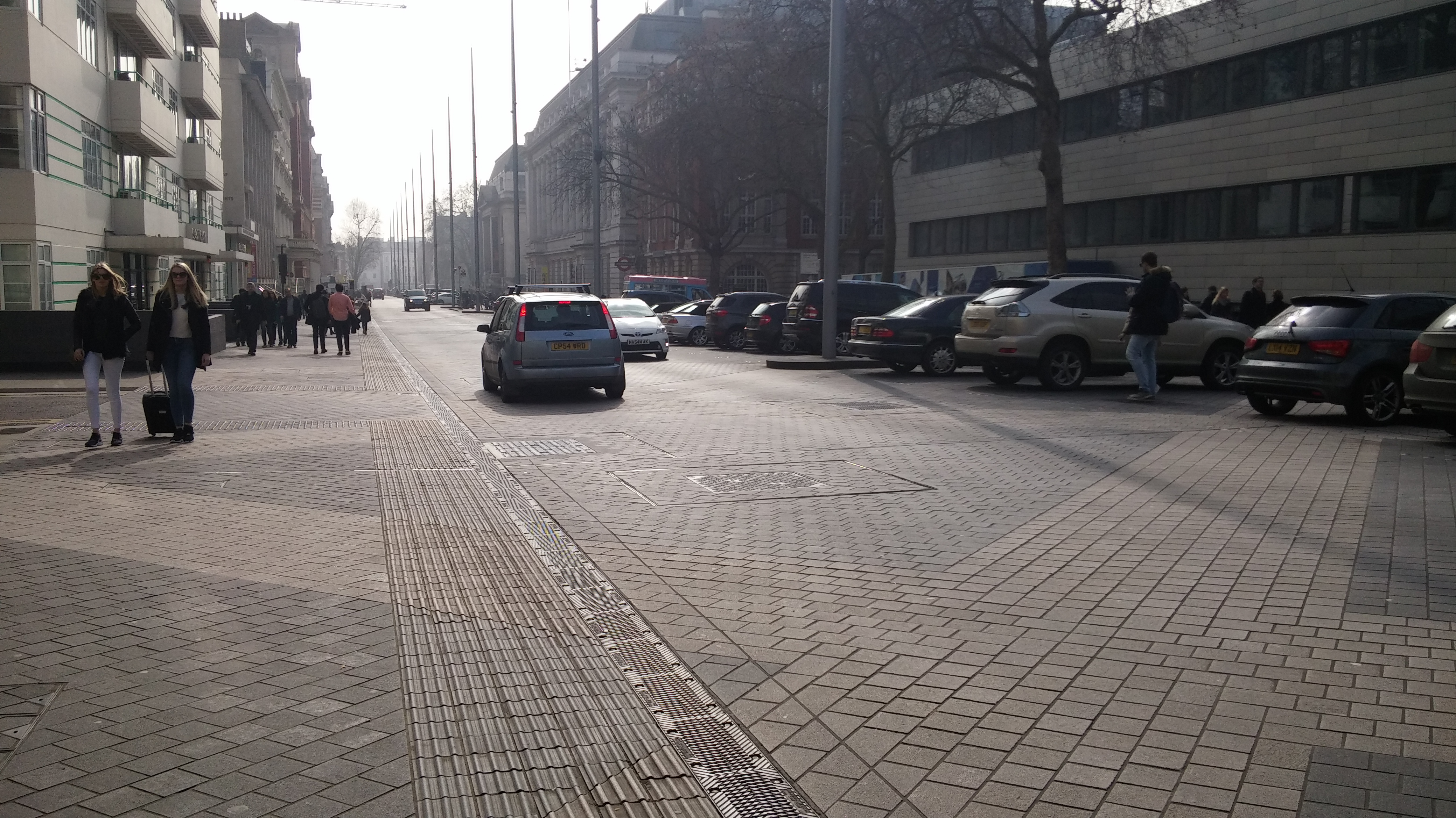 A wide paved surface between two building lines with a criss-cross tiled pattern. Parked cars to the right hand side. Cars drive down the middle. A small amount of tactile paving and a drainage grille marks out the central area where cars drive. People walk down the left hand side towards the camera. There are pedestrians and cars moving in the same space with no physical separation.